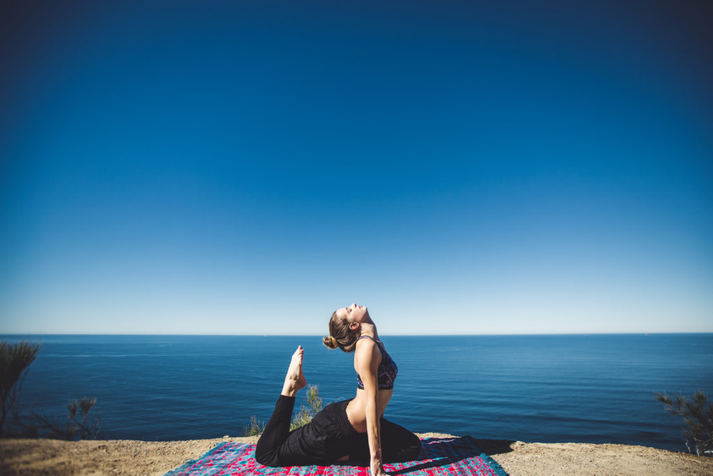  serene outdoor yoga scene where someone is performing dhanurasana (bow pose) on a yoga mat. The setting shows a beautiful ocean view with a clear blue sky meeting the water at the horizon.