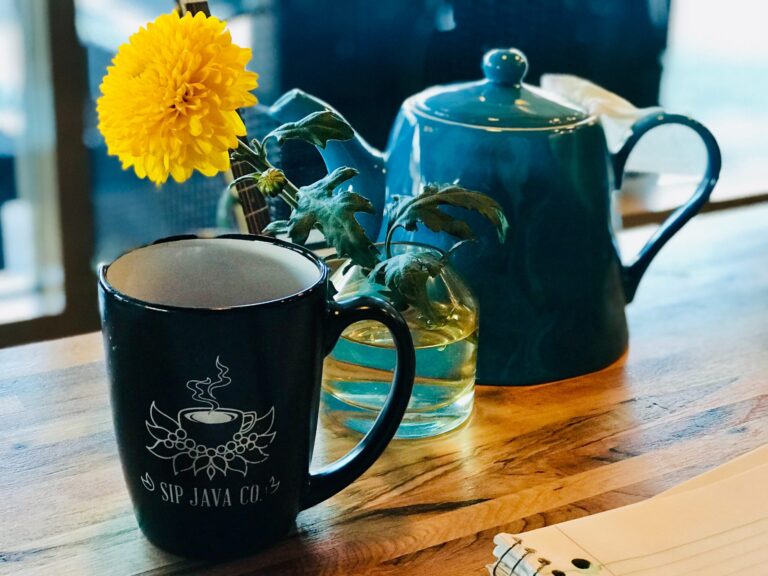 The image shows a cozy coffee shop scene with a dark ceramic mug featuring the "Sip Java Co" logo, a teapot, and a yellow chrysanthemum flower against a warm, wooden background. The scene has a calm, inviting atmosphere.