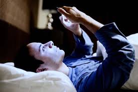 Person lying in bed in a dark room, illuminated by the blue light from a smartphone or tablet they are holding above their face while using it.