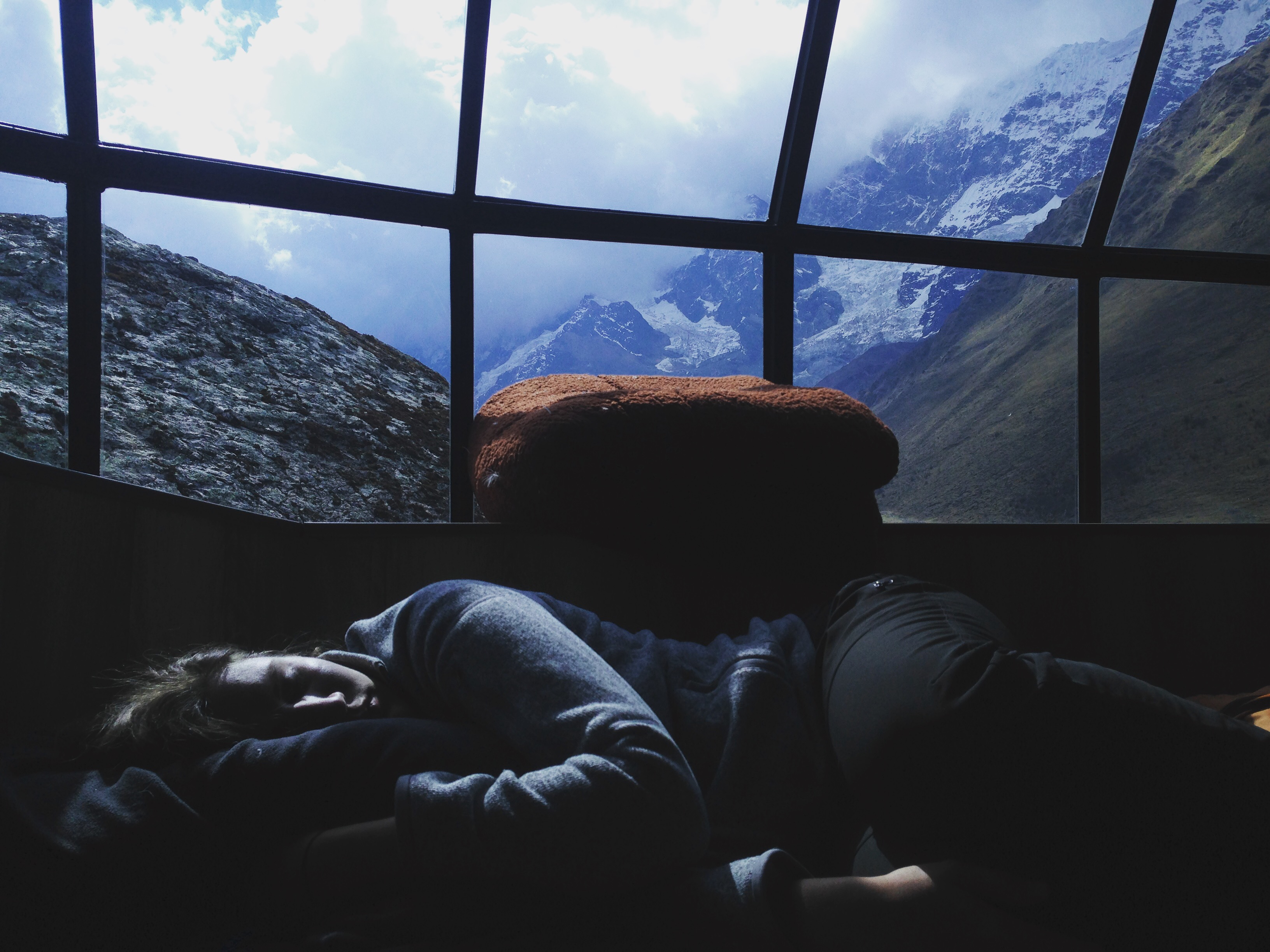 Person sleeping on a couch or window seat with a panoramic view of snow-capped mountains through large windows. The scene contrasts the cozy interior with the dramatic mountain landscape outside.