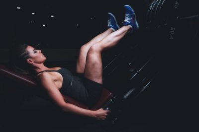 Person performing a leg press exercise on a machine, wearing blue sneakers and a black athletic outfit, in a dimly lit gym