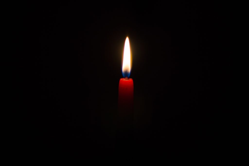 Shows a single red candle lit against a completely black background. The flame glows brightly in the darkness, creating a strong visual contrast
