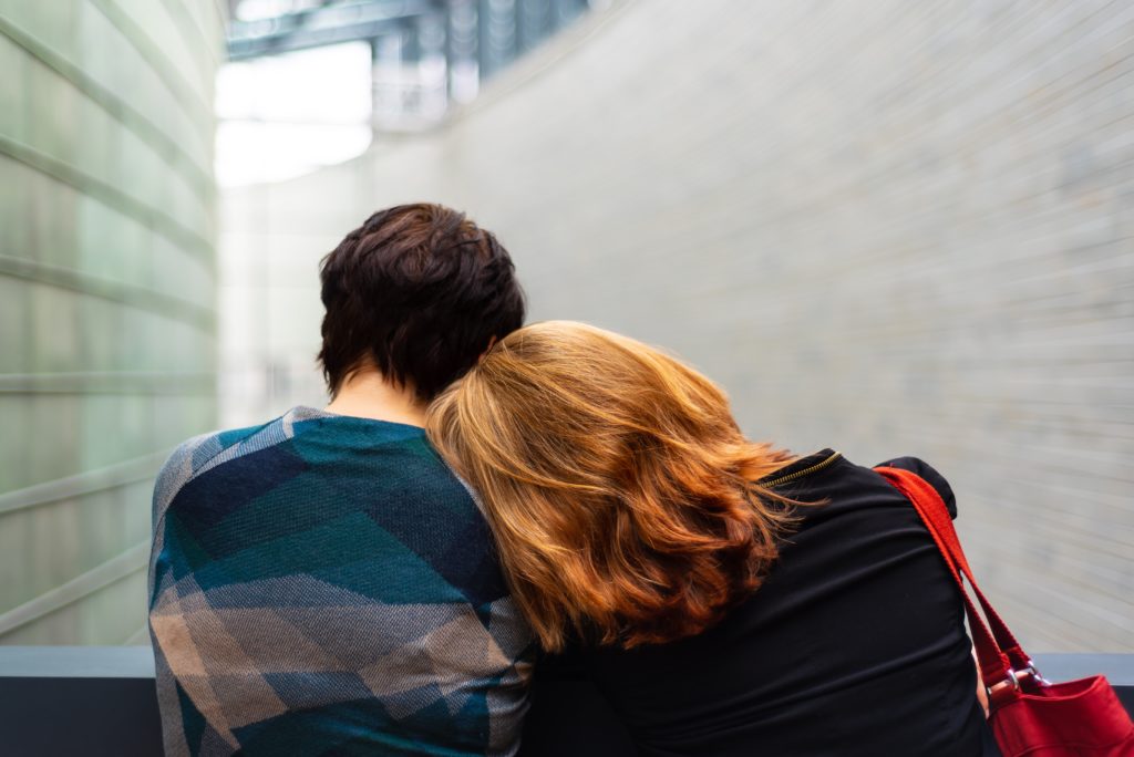  Shows two people from behind, sitting close together. One person has short dark hair and is wearing a blue patterned garment, while the other has shoulder-length reddish/auburn hair and is leaning their head on the first person's shoulder. They appear to be in what looks like a modern building with light-colored walls. A red bag or item is visible to the side. The image conveys intimacy and emotional connection.