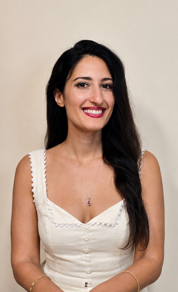 Smiling woman with long dark hair wearing a white dress and delicate gold jewelry, photographed against a neutral beige background for a personal brand headshot.